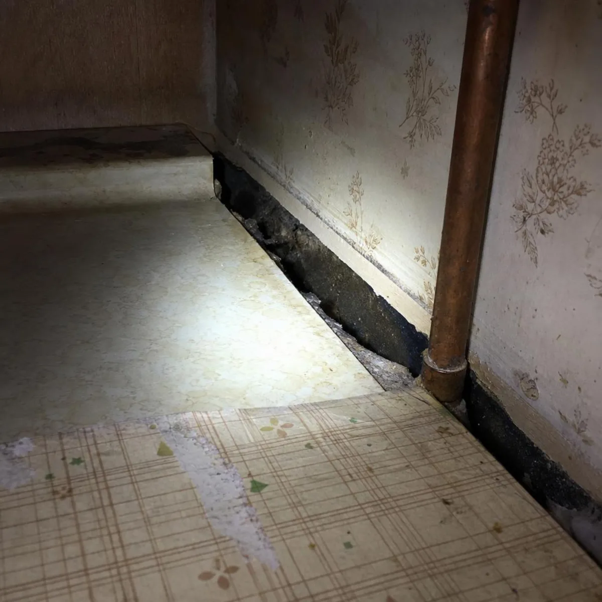 Inside a dim apartment kitchen cabinet, a gap runs along the seam where the counter meets the wall next to a plumbing pipe.