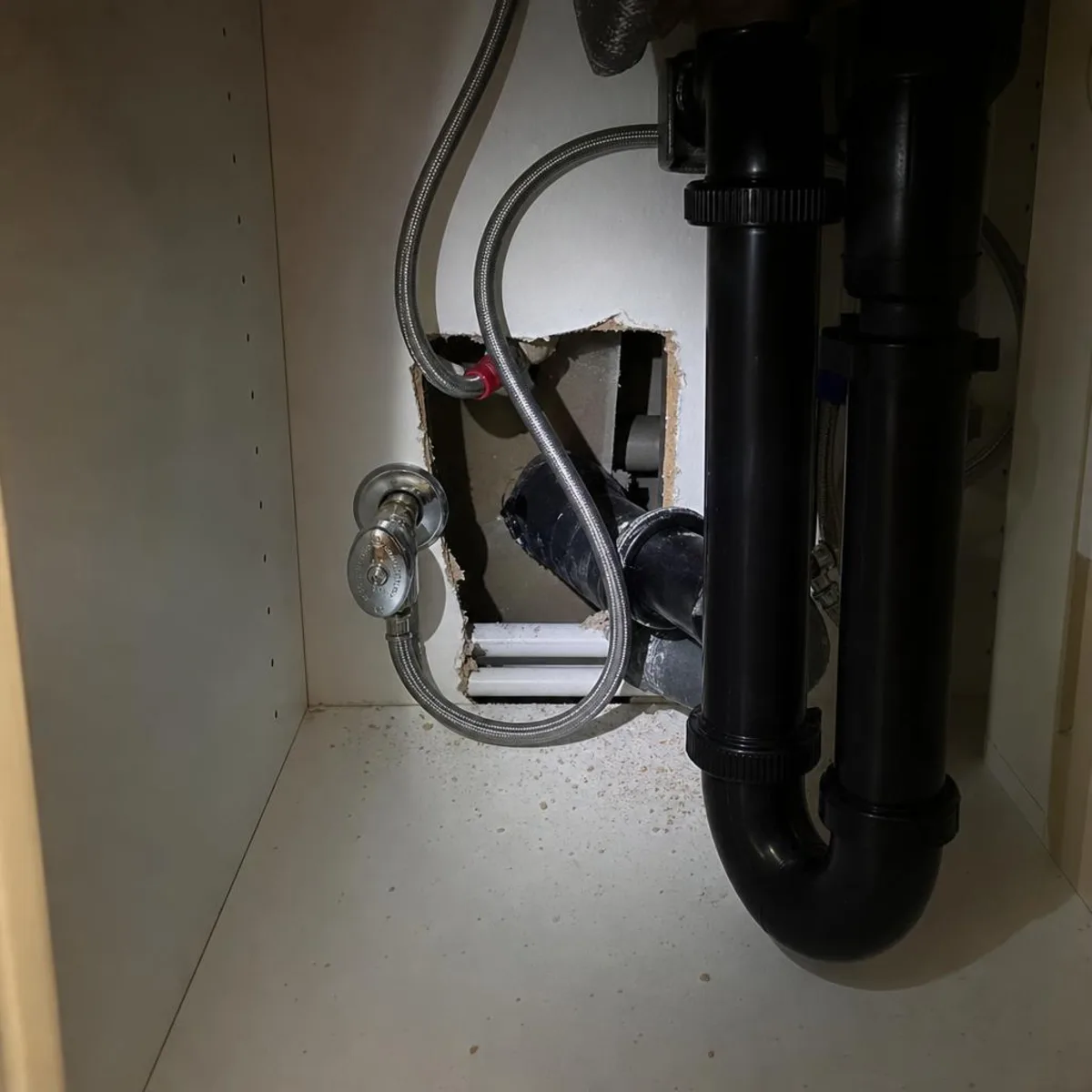 The inside of a condominium kitchen sink cabinet, where the drain trap and supply lines pass through the back wall toward the neighbouring unit.