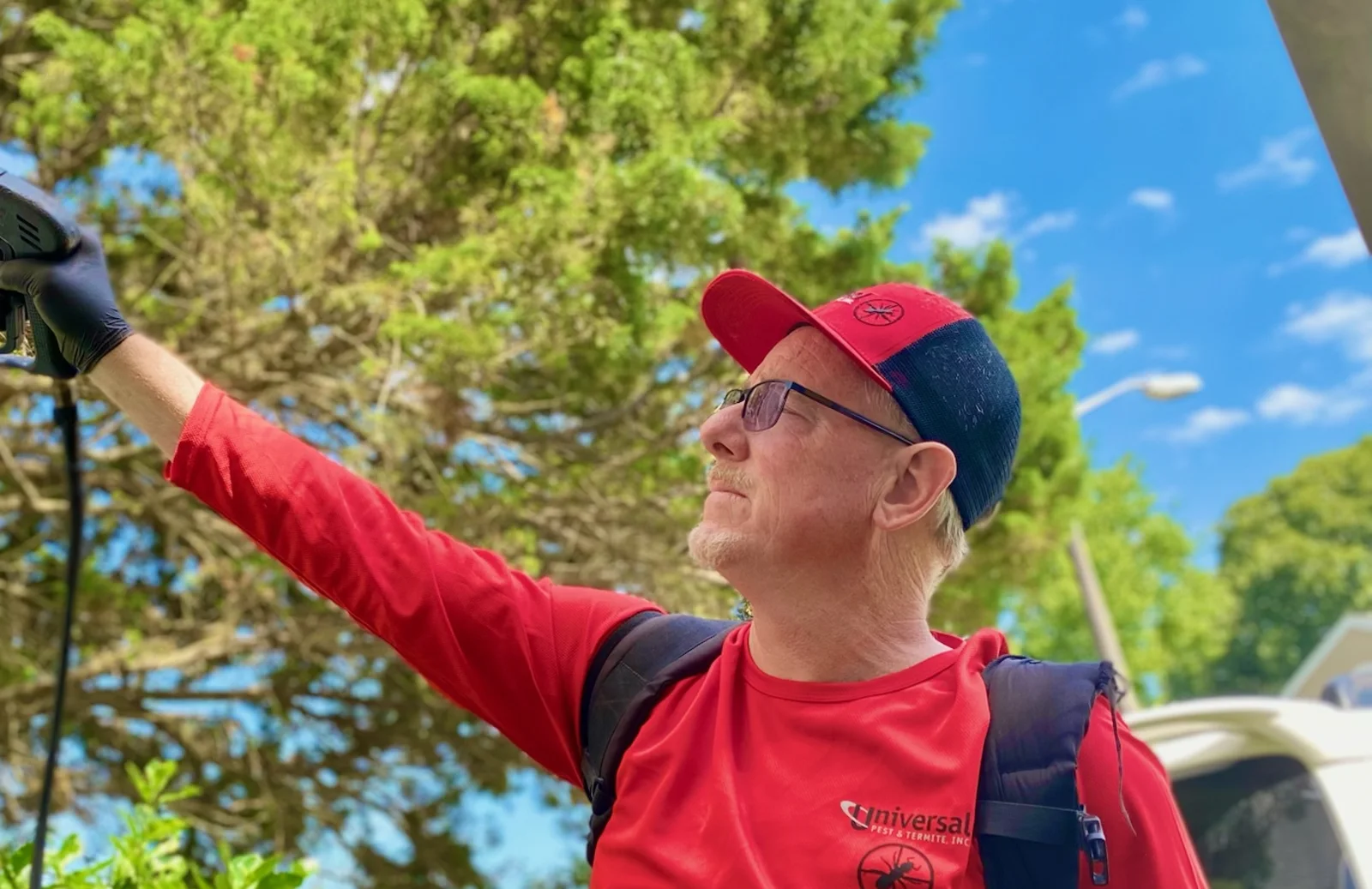 A Universal Pest and Termite technician in a red long-sleeve uniform shirt and red and navy cap raising a backpack sprayer wand toward the eaves of a home, the service van parked behind him under a clear blue sky.
