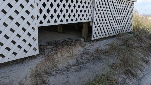 Wooden lattice skirting around the crawl space of a raised beach cottage, with sandy soil washed away below it leaving an open gap.