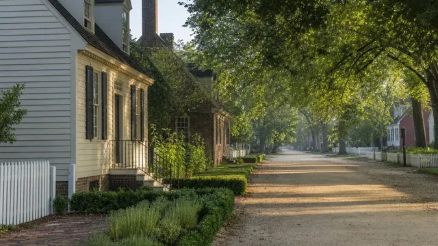 A tree-shaded colonial street in Williamsburg at first light, with white clapboard and brick houses behind picket fences, brick walkways and clipped boxwood hedges beside a sandy unpaved lane.