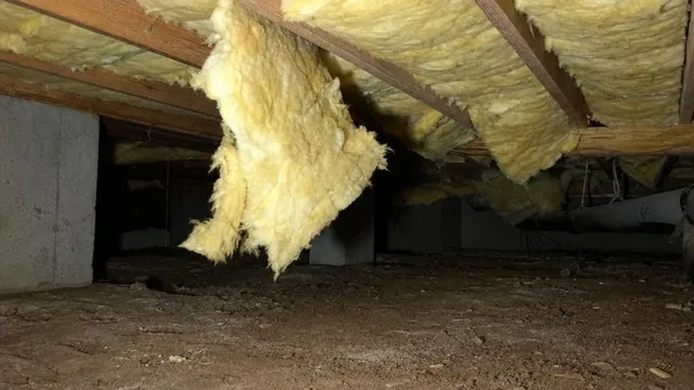 Inside a crawl space, fiberglass insulation batts sag away from the floor joists above bare soil, with one section pulled loose and hanging down.