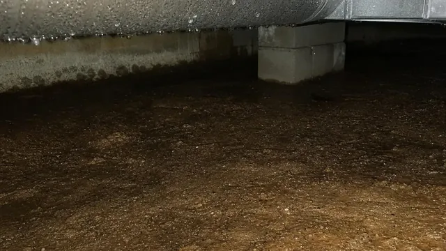 A metal duct running across a crawl space above dark, damp bare earth, with moisture staining in the soil beneath the run.