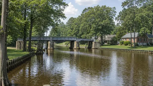 The Albemarle and Chesapeake Canal at Great Bridge on a summer morning, with a steel drawbridge crossing the still water, bald cypress along the near bank and brick homes with mown lawns on the far side.