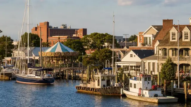 Sailboats and workboats moored along the downtown Hampton waterfront at sunset, the historic carousel pavilion and low brick buildings lining the harbor walk beside the calm Hampton River.