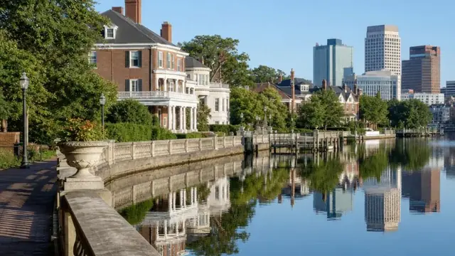 Historic brick and stucco homes of Ghent lining the curved seawall of The Hague inlet in Norfolk at golden hour, sailboats moored on the calm water and the downtown skyline rising in the distance.