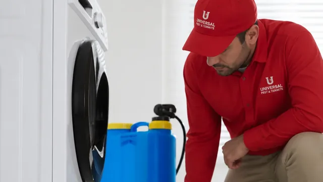 Universal Pest & Termite technician in a red branded shirt and cap kneeling beside a washing machine during an indoor pest treatment.