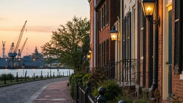 Brick rowhouses with wrought-iron fences and gas lamps along a tree-shaded Olde Towne Portsmouth street in the late afternoon, shipyard cranes visible over the rooftops toward the Elizabeth River.