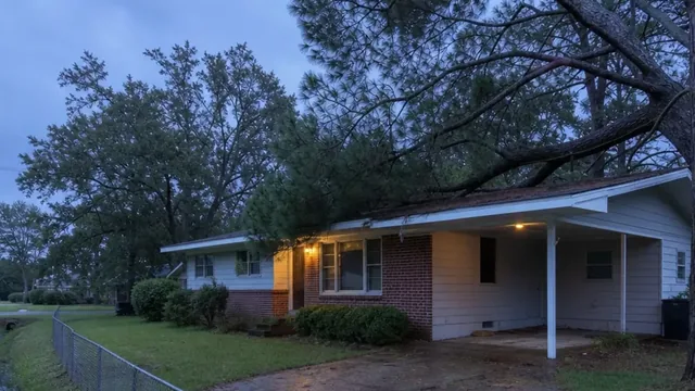 A low-pitched 1960s brick and siding ranch house with a carport at dusk, heavy oak and pine branches resting on the roof edge and gutter, and a grassy drainage ditch behind a chain-link fence at the edge of the yard.