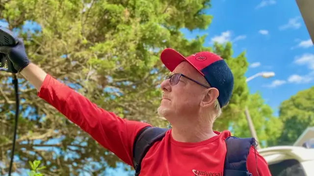 A Universal Pest and Termite technician in a red long-sleeve uniform shirt and red and navy cap raising a backpack sprayer wand toward the eaves of a home, the service van parked behind him under a clear blue sky.