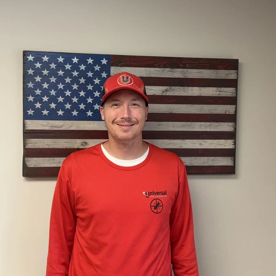 Brandon Nikolaisen, Manager at Universal Pest & Termite, wearing a red company shirt and cap in front of an American flag