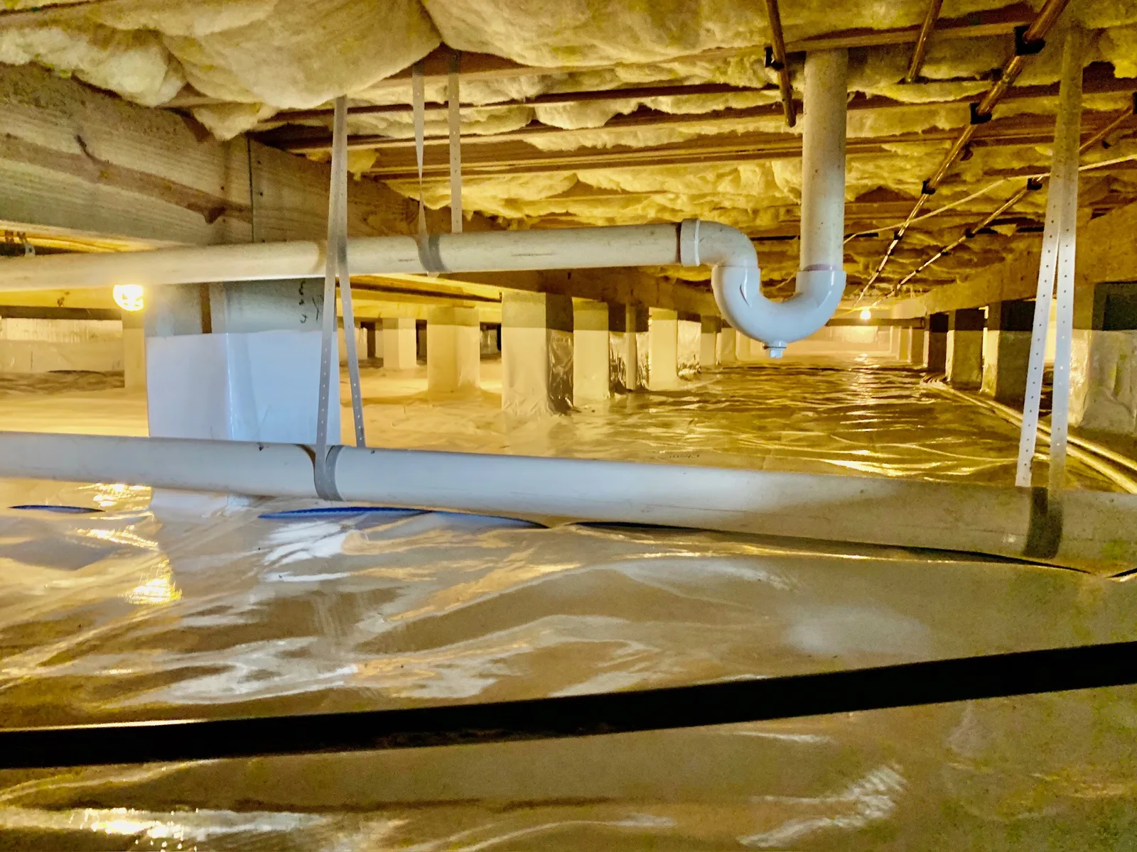 Encapsulated Hampton Roads crawl space with a reinforced vapor barrier sealed across the ground and up the piers, insulated joists overhead, and lit, dry access between the drain lines
