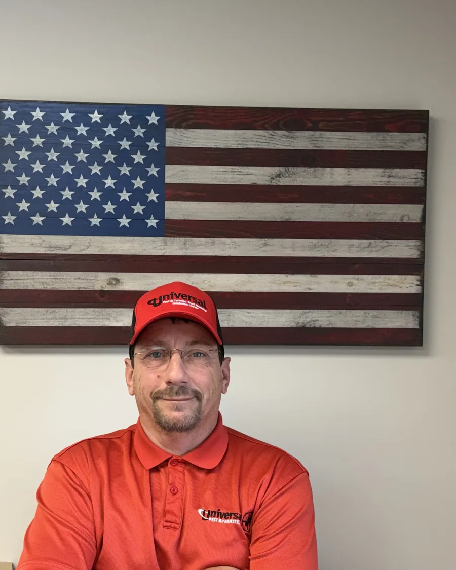 Jimmy, a Virginia State Certified pest control professional with Universal Pest & Termite, wearing a red company polo and cap in front of a wooden American flag