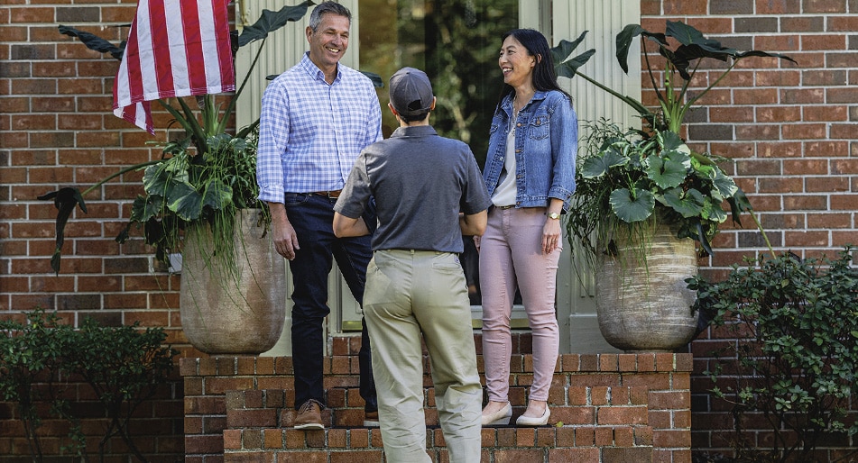 A man and woman stand on the steps of a house, proudly displaying an American flag in the background.